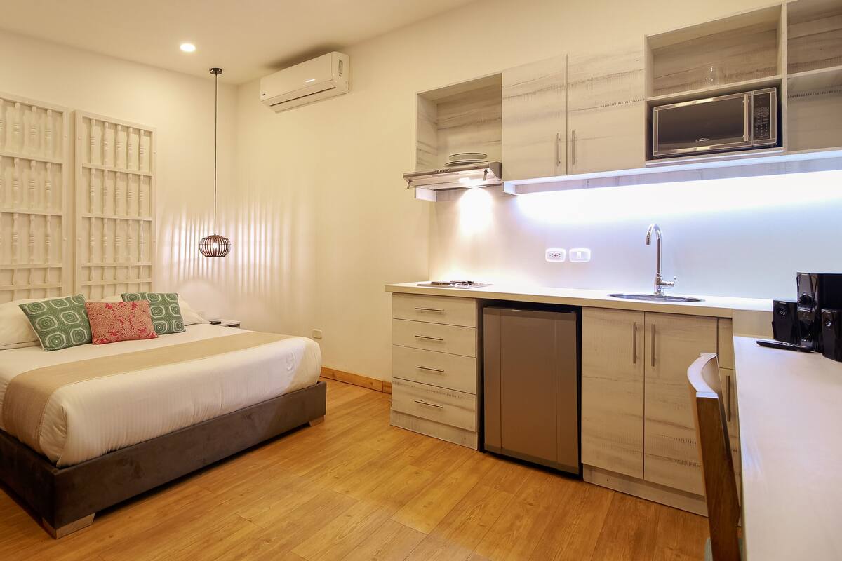 Una habitación compacta con cama, mesitas de noche, armario empotrado y una pequeña cocina con microondas y fregadero.