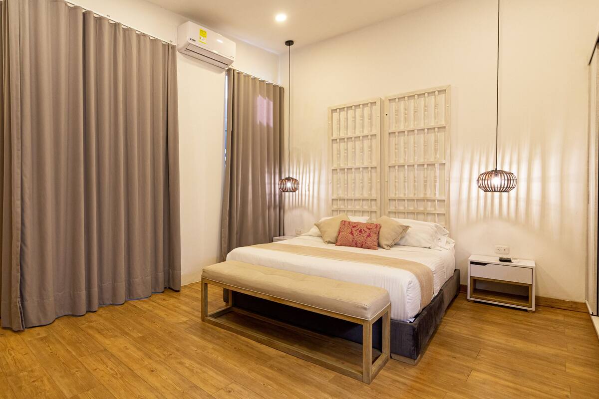 Un dormitorio con cama, mesita de noche, banco, cortinas y un equipo de aire acondicionado.