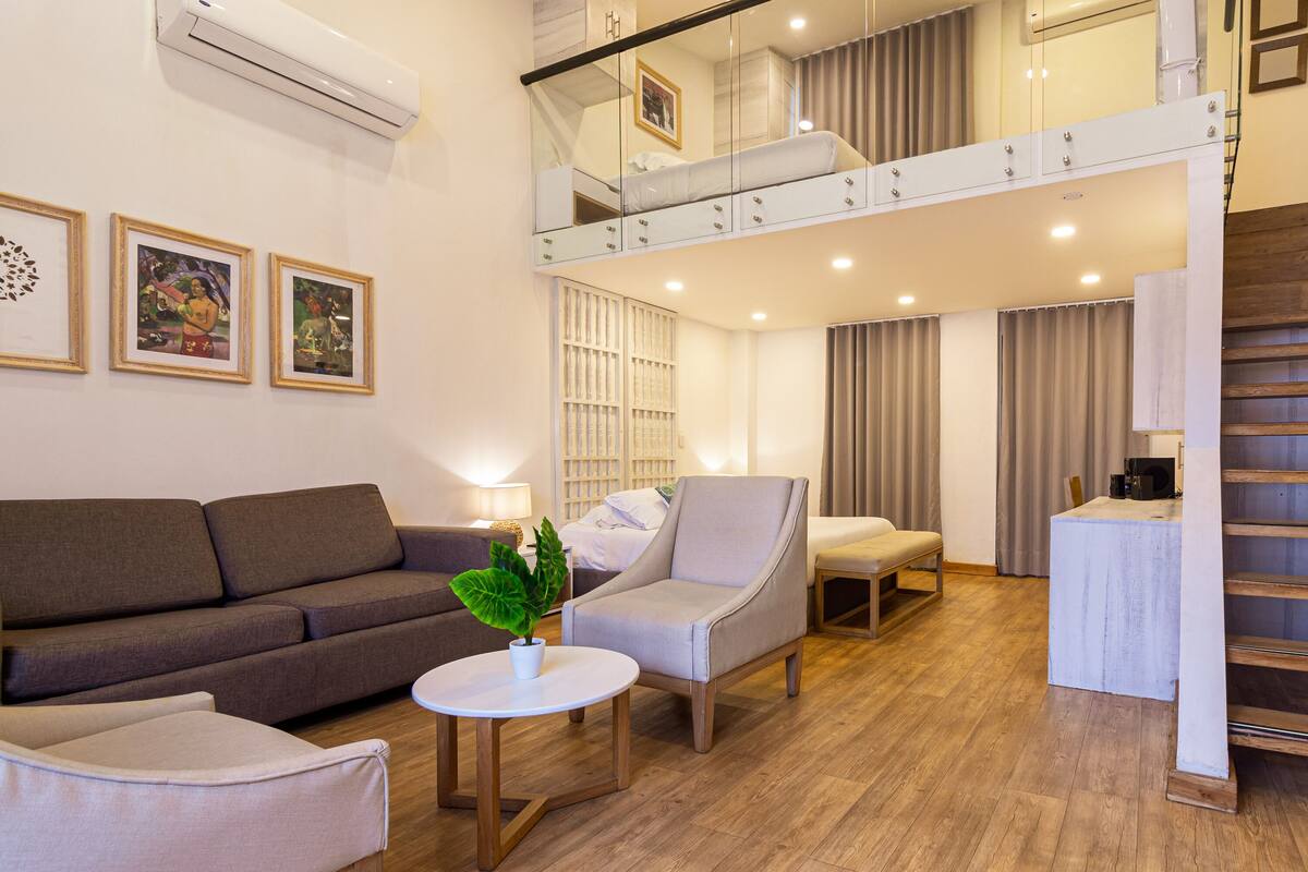 Una habitación de hotel moderna con un sofá, sillones, una mesita, una escalera y un balcón.
