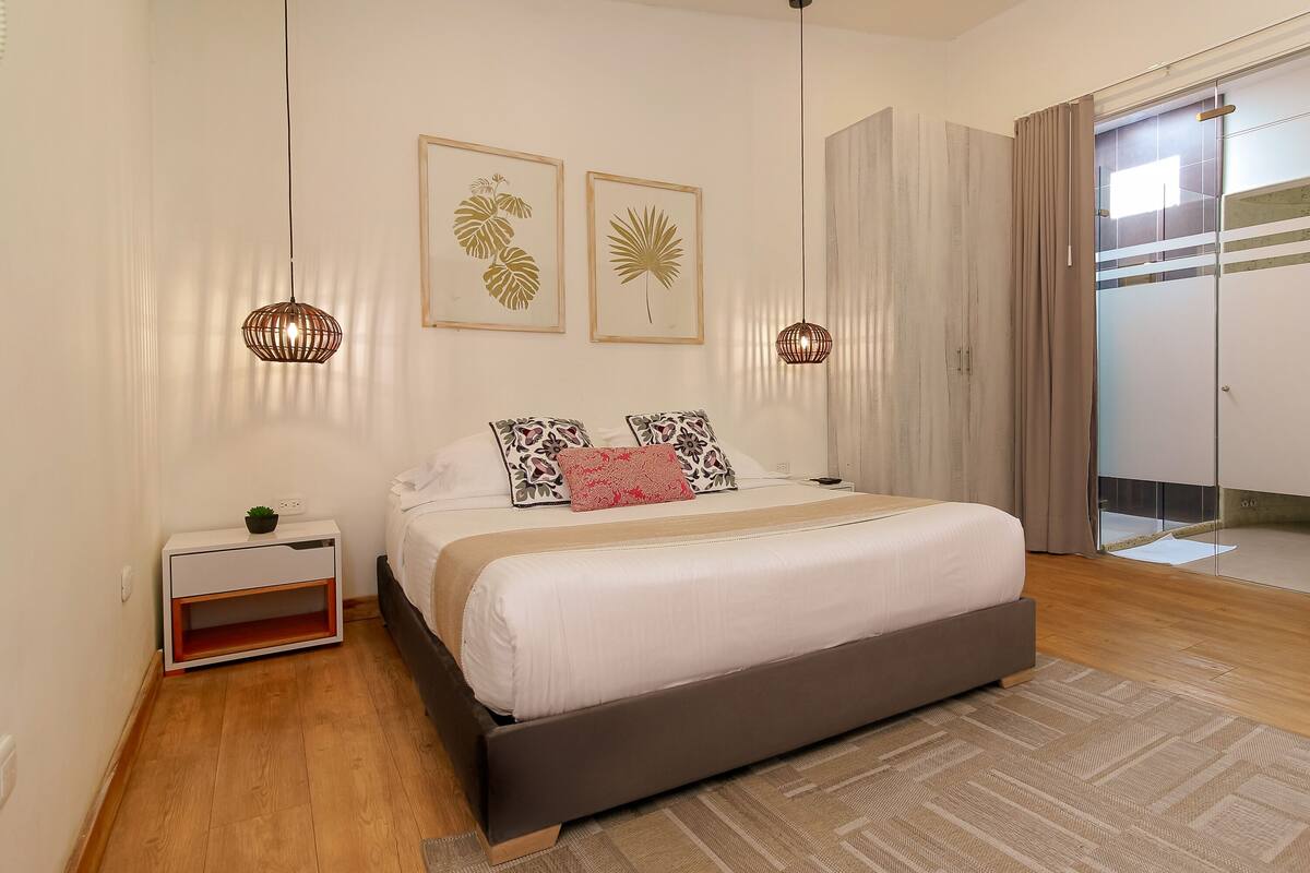 Un dormitorio moderno con una cama grande, mesitas de noche, una planta pequeña y dos luces fijadas a la pared.