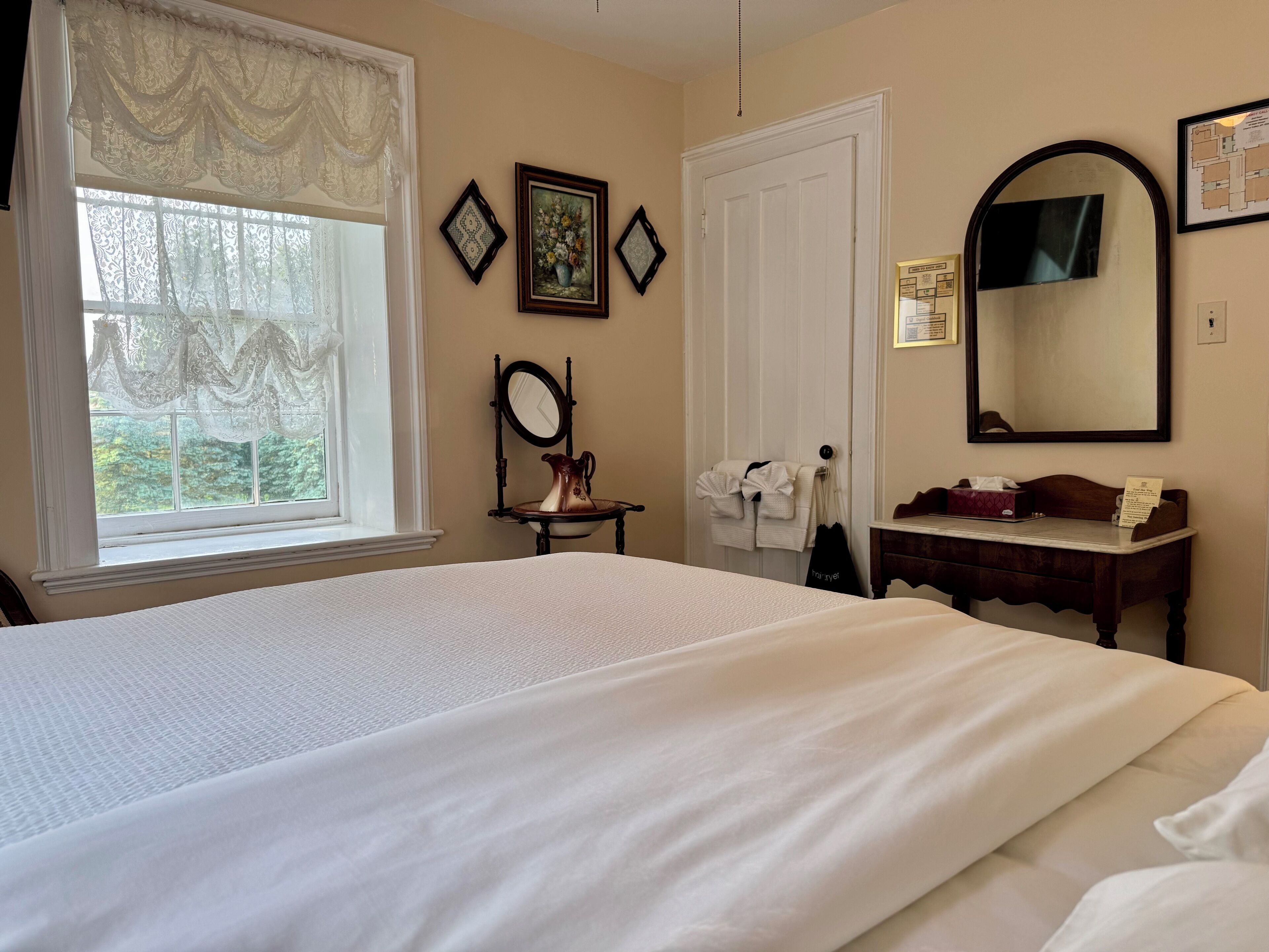 Ein Hotelzimmer mit einem großen Bett, einem Schminktisch, einem Spiegel und einem Fenster mit Spitzenvorhängen.