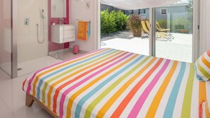 A bedroom with a colorful striped bedspread, a white vanity with a mirror, a shower area, and a view of a garden with outdoor seating.