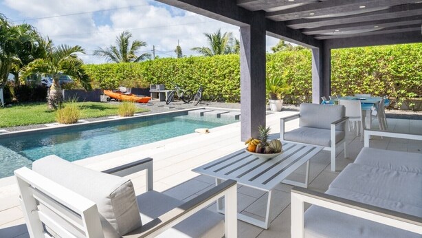 A covered outdoor area with a pool, white furniture, and a view of a lush garden.
