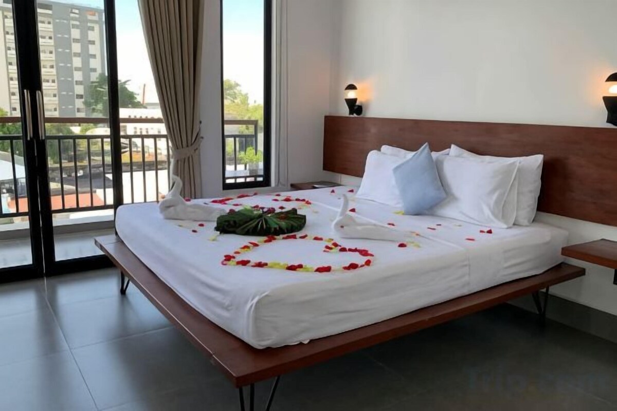 Een hotelkamer met een bed, nachtkastje, raam met gordijnen en uitzicht op gebouwen.