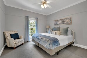 Una camera da letto con un letto, una sedia, una ventola a soffitto, una finestra con tende e un cartello appeso al muro.