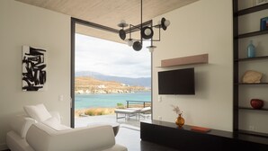 Ein modernes Wohnzimmer mit einem weißen Sofa, einer schwarzen Unterhaltungsanlage und einem großen Fernseher an der Wand. Durch eine Glastür ist der Blick auf das Meer.