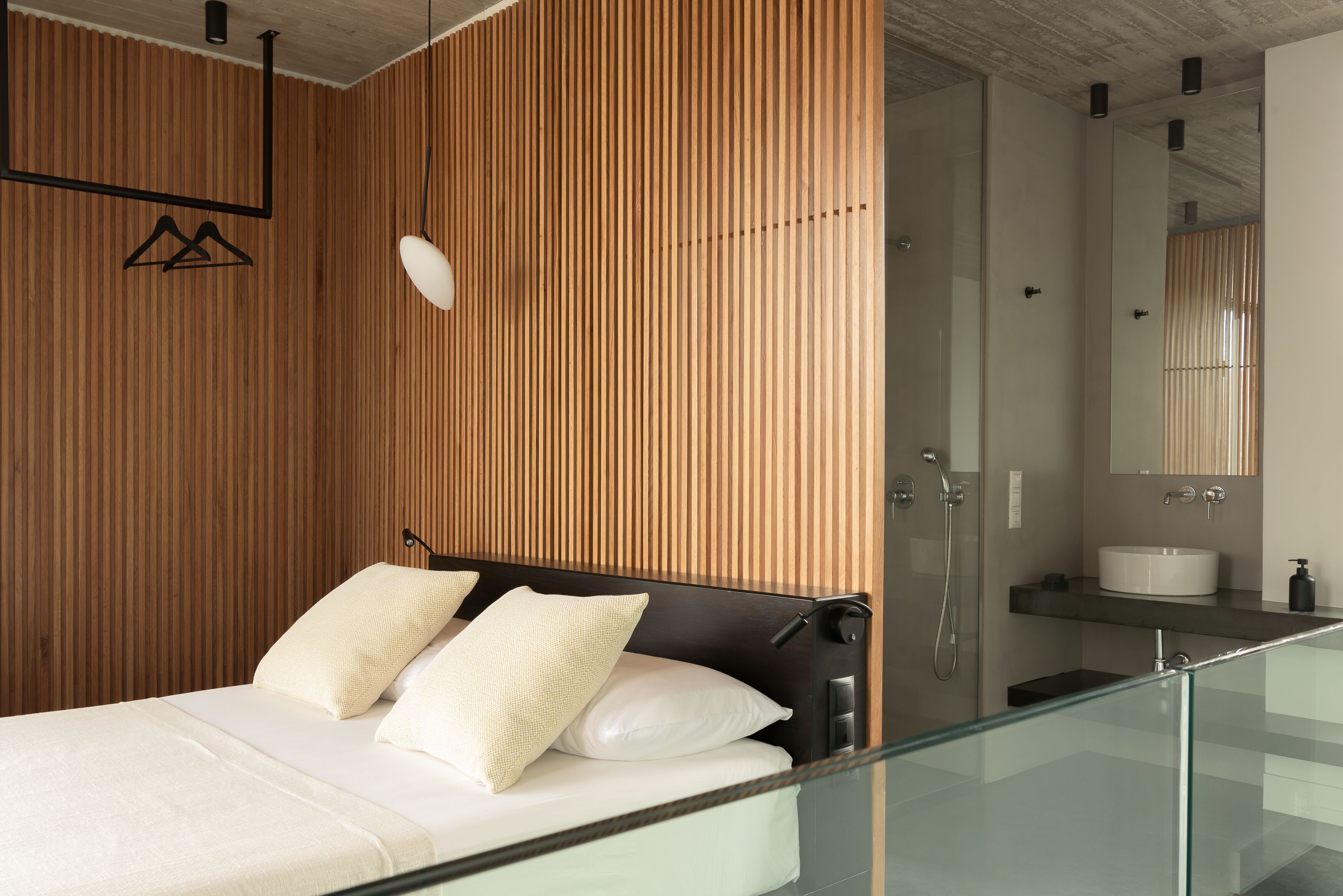Un dormitorio moderno con pared de paneles de madera, balcón de vidrio, cama con ropa de cama blanca y baño con lavamanos y espejo blancos.