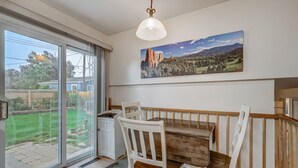 A dining area with a wooden table and white chairs, a large window with a view of a backyard, and a painting of a landscape on the wall. - Sherwood (Colorado Springs)