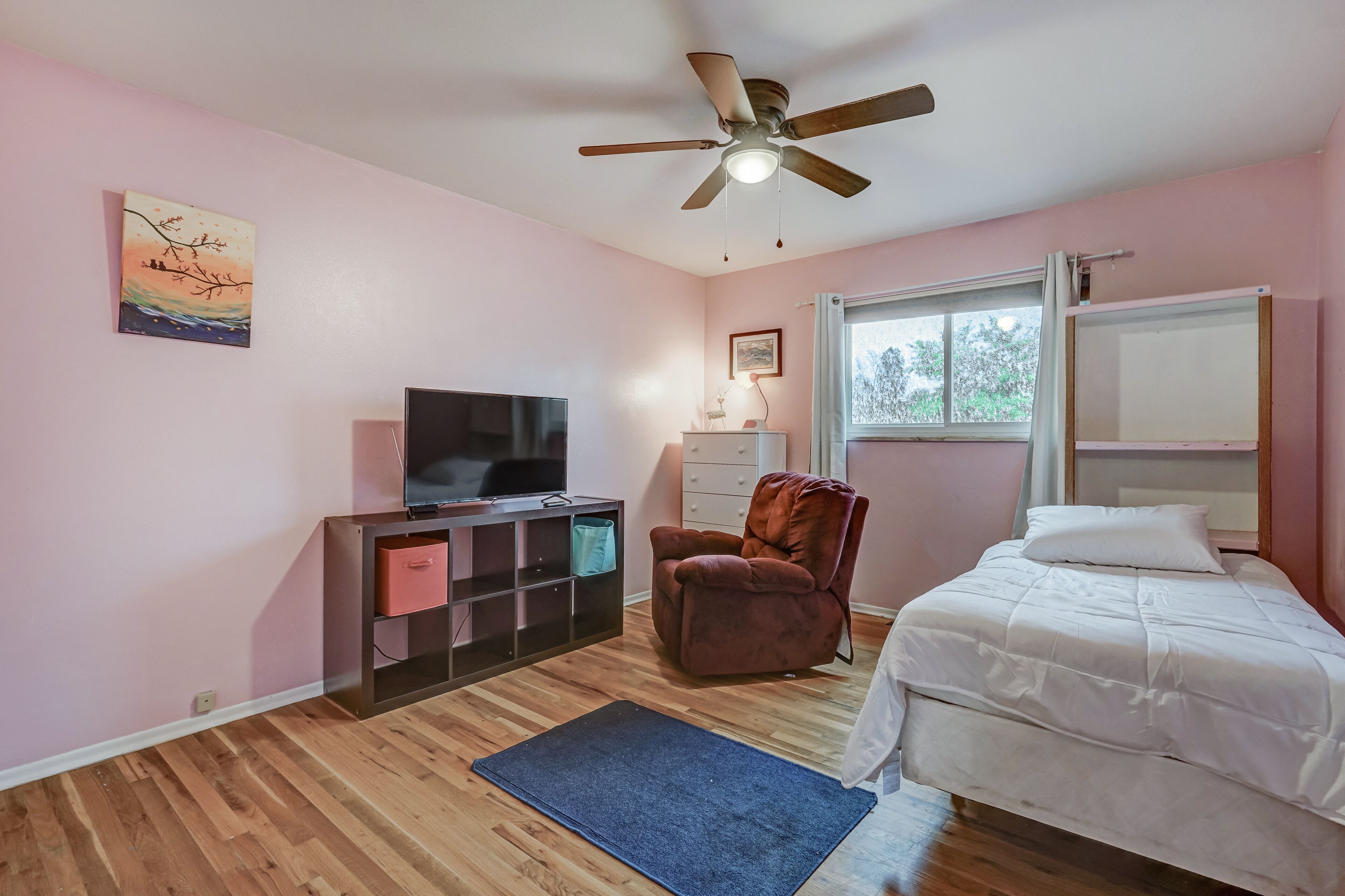 A bedroom with a bed, a television, a chair, a ceiling fan, and a window.