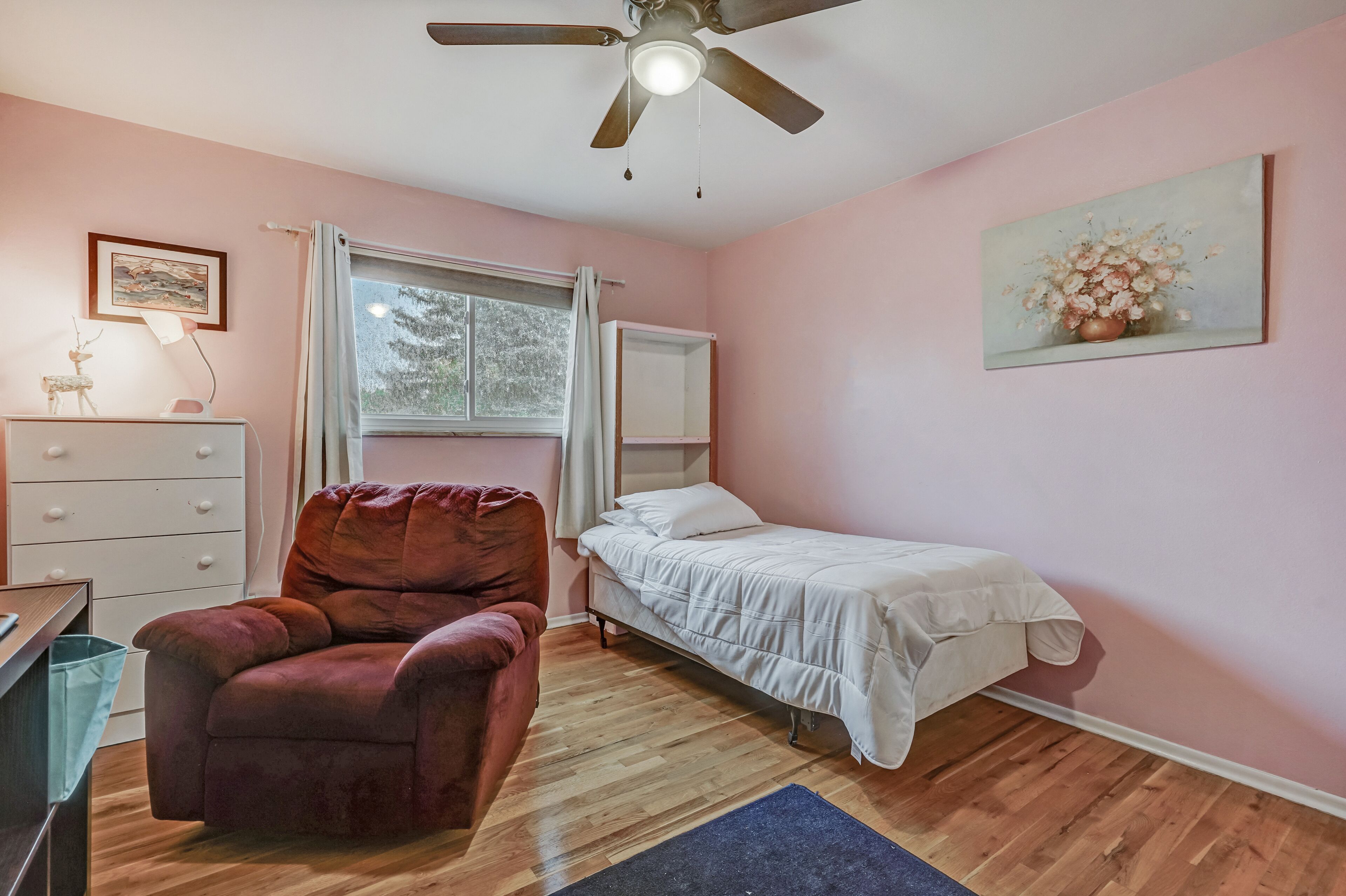 A bedroom with a single bed, a chair, a desk, a ceiling fan, and a window with curtains.