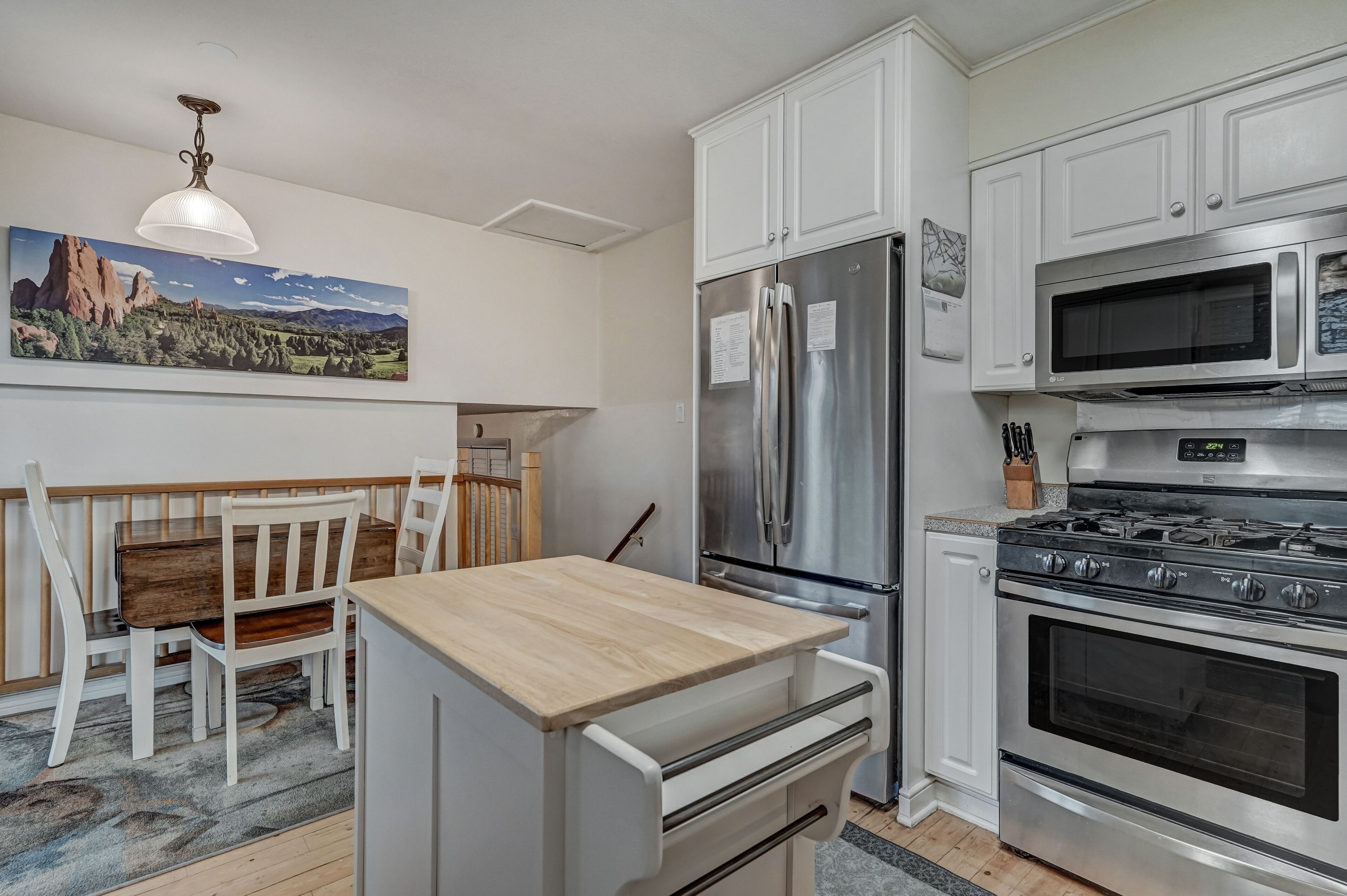 A modern kitchen with white cabinets, stainless steel appliances, a wooden dining table, and a large landscape painting on the wall.