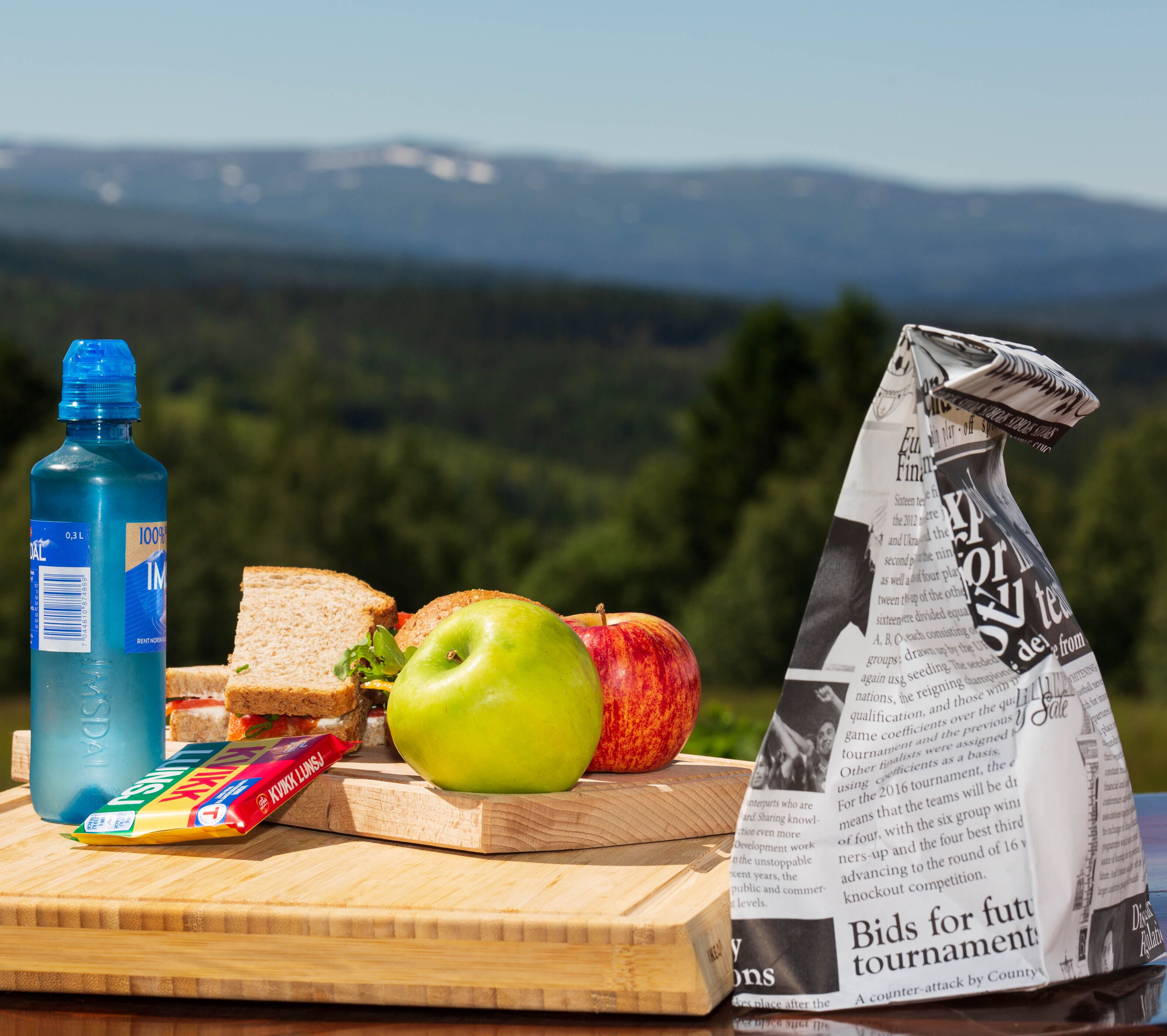 A meal with a sandwich, apple, juice bottle, and newspaper on a wooden table with a scenic mountain view in the background.