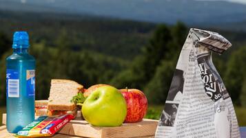 A meal with a sandwich, apple, juice bottle, and newspaper on a wooden table with a scenic mountain view in the background.