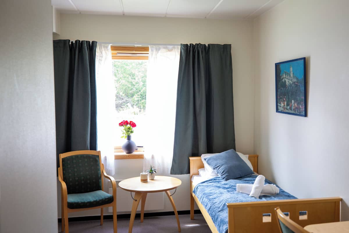 Ein Zimmer mit einem Bett, einem Sessel, einem kleinen Tisch und einem Fenster mit Vorhängen.