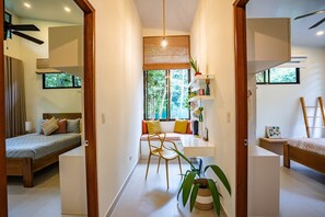 A modern, well-lit room with a bed, a desk, and a chair. There is a large window providing natural light and a view of greenery outside.
