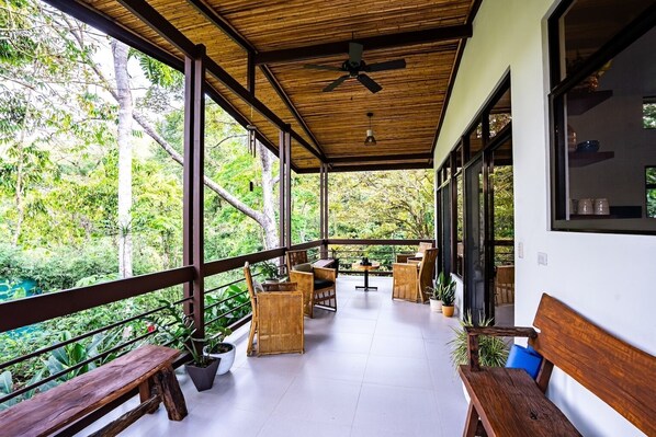 A covered outdoor area with wooden furniture and potted plants, overlooking a lush green landscape.
