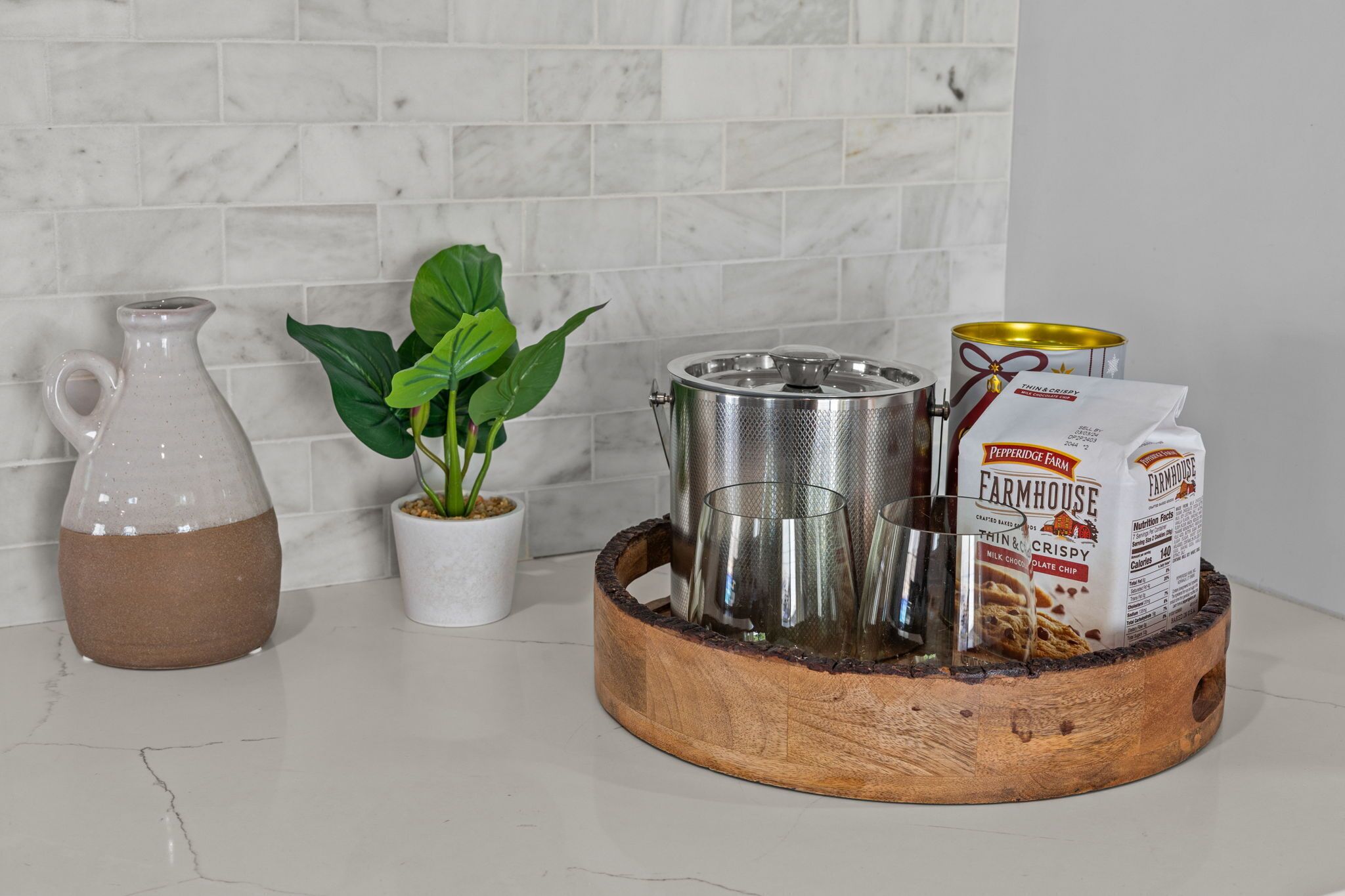 A kitchen counter with a marble backsplash, a ceramic pitcher, a potted plant, a metal pot, a wooden tray, and a can of milk.