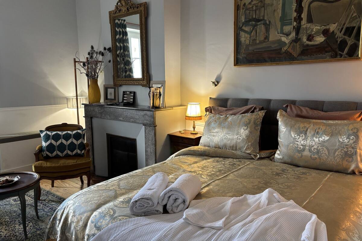 A hotel room with a large bed, a fireplace, a nightstand with a lamp, a chair, and a painting on the wall.