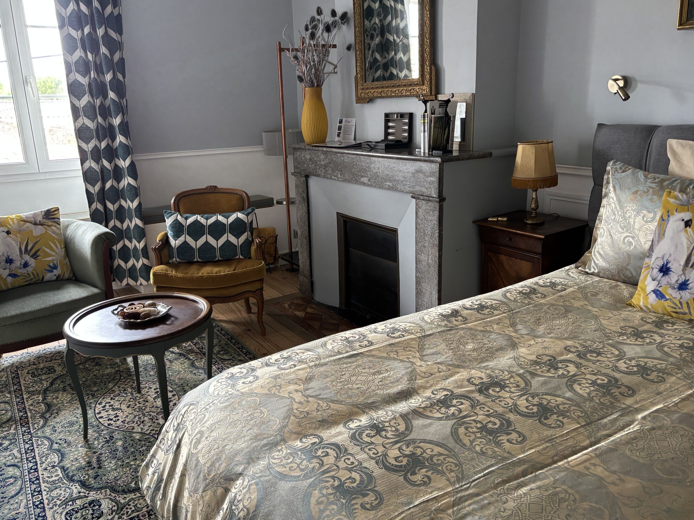 A room with a bed, a sofa, a chair, a small table, a fireplace, and a mirror.