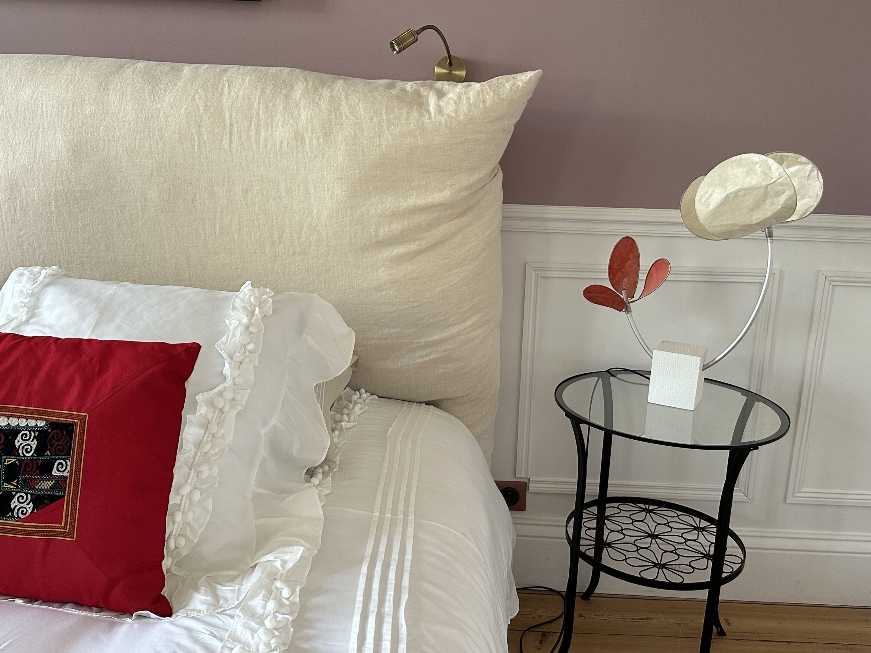 Una cama con un almohadón blanco, un cojín decorativo rojo y una mesita redonda con una pantalla blanca.