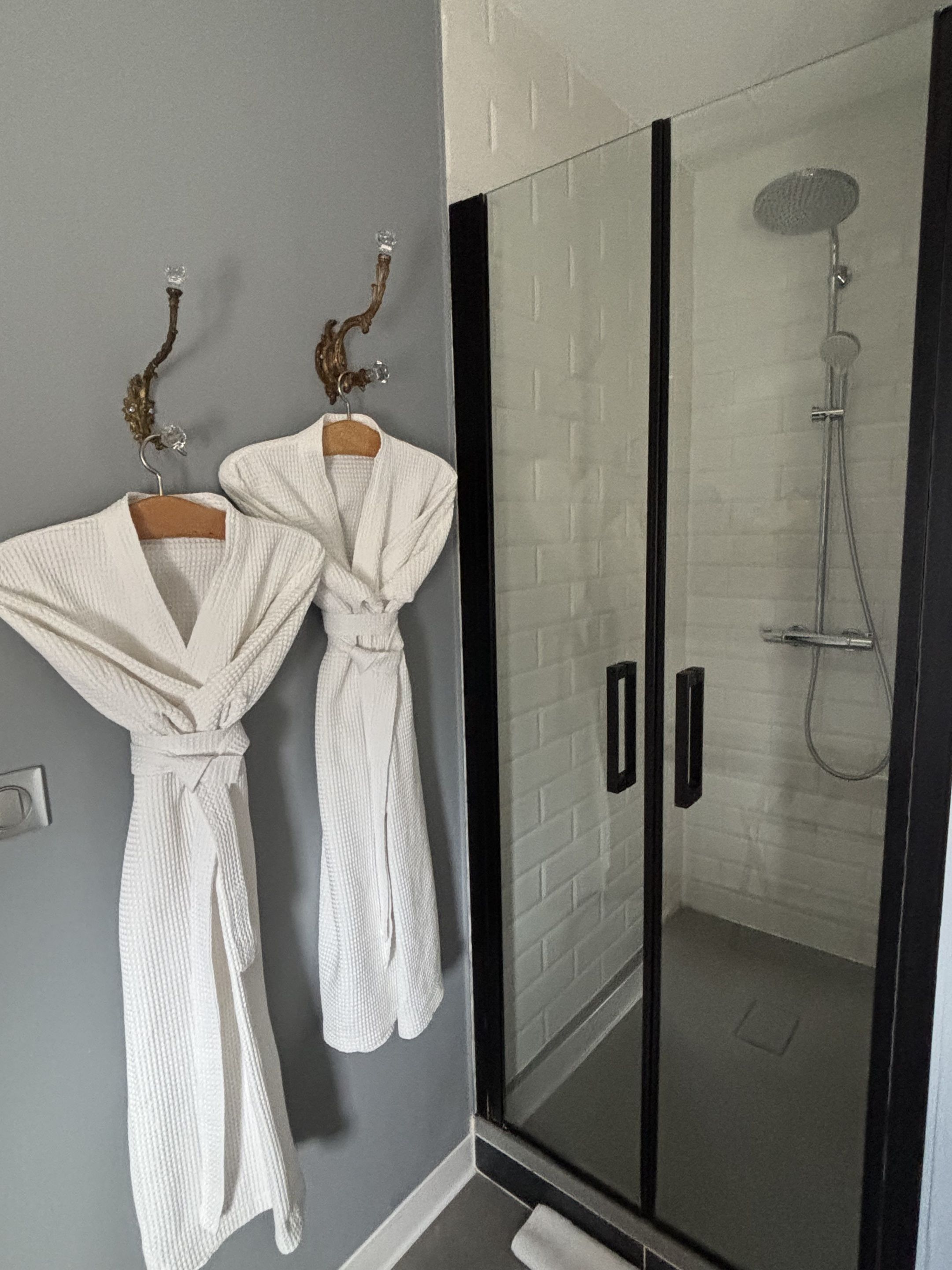 Een badkamer met twee witte badjassen aan een muur, een douchecabine van glas en een handdoekhouder met een decoratief object.