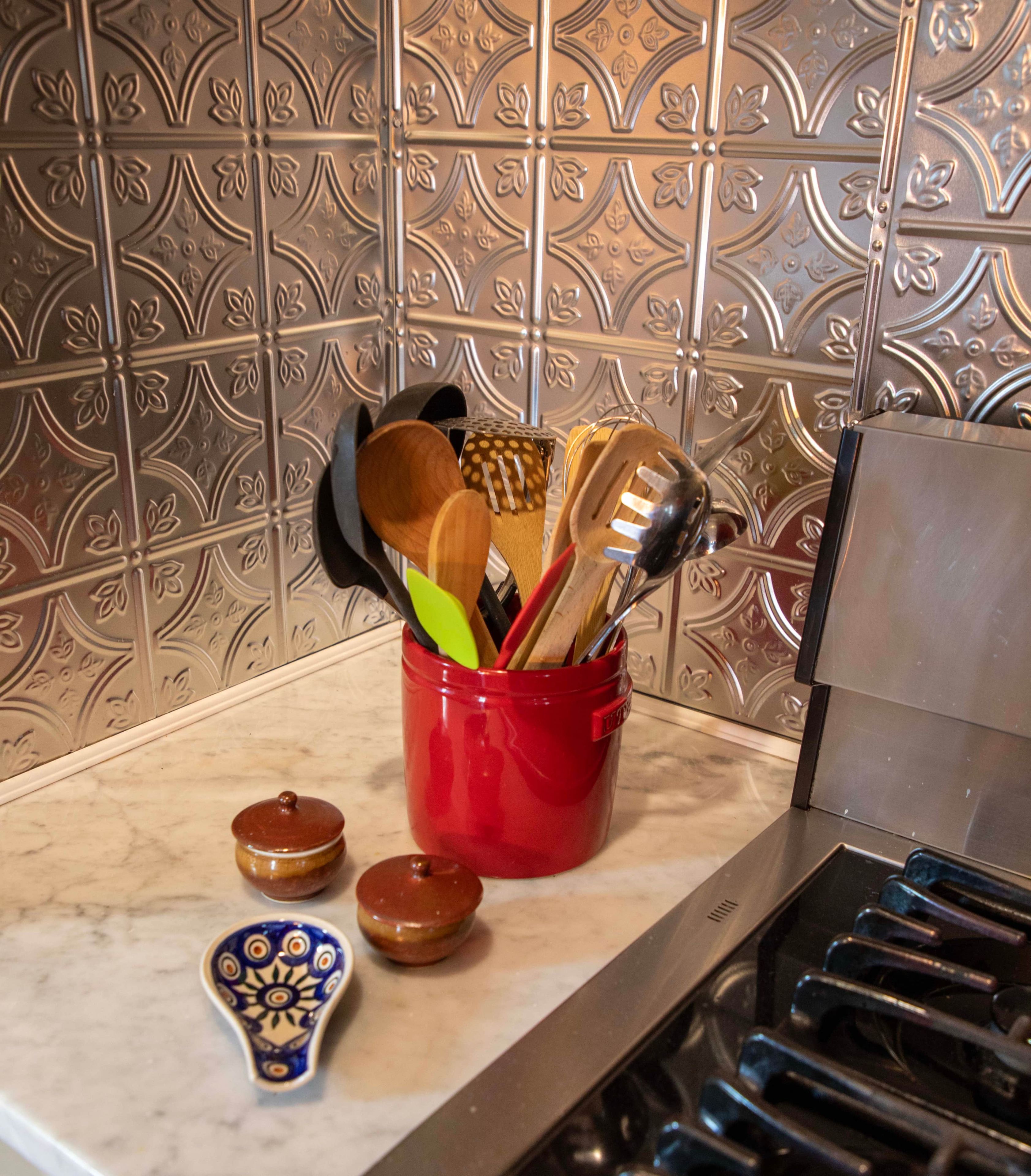 Uma bancada de cozinha com um porta-utensílios vermelho contendo utensílios de madeira e plástico, uma colher de cerâmica azul e branca, e dois pequenos potes com tampas.