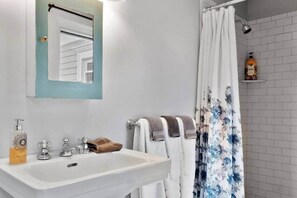 Un bany amb un vàter blanc, dues aixetes, un mirall amb marc blau, una cortina de bany amb dibuixos florals i una ampolla de xampú sobre una prestatgeria.