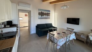 En kompakt opholdsstue med tekøkken, spiseplads med bord og stole, sofa og tv ophængt på væggen.