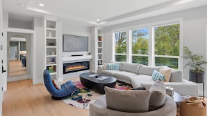 A modern living room with a fireplace, a sofa, a coffee table, and a large window with a view of trees. - Luxury 3 King Suite Retreat 3 Blocks to Downtown! (Carlton)