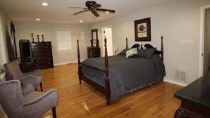 Een slaapkamer met een hemelbed, een plafondventilator, houten vloeren, een dressoir, een stoel en een ingelijst schilderij aan de muur.