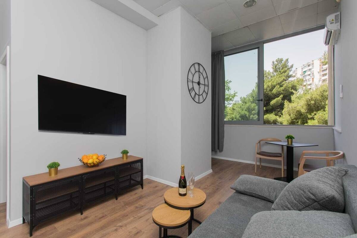 A modern living room with a flat-screen TV, a wooden console table with fruit, a round table with a bottle, and a grey sofa.