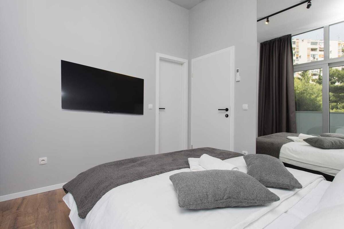 A modern bedroom with a large bed, a smaller bed, a flat-screen TV, and a balcony with curtains.