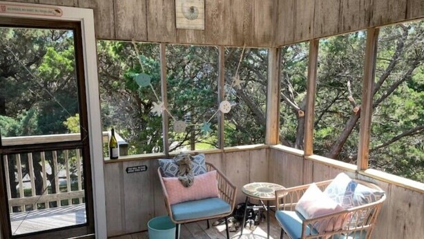 Una terraza de madera con mobiliario de mimbre y una pequeña mesa, rodeada de grandes ventanales con vista a árboles.