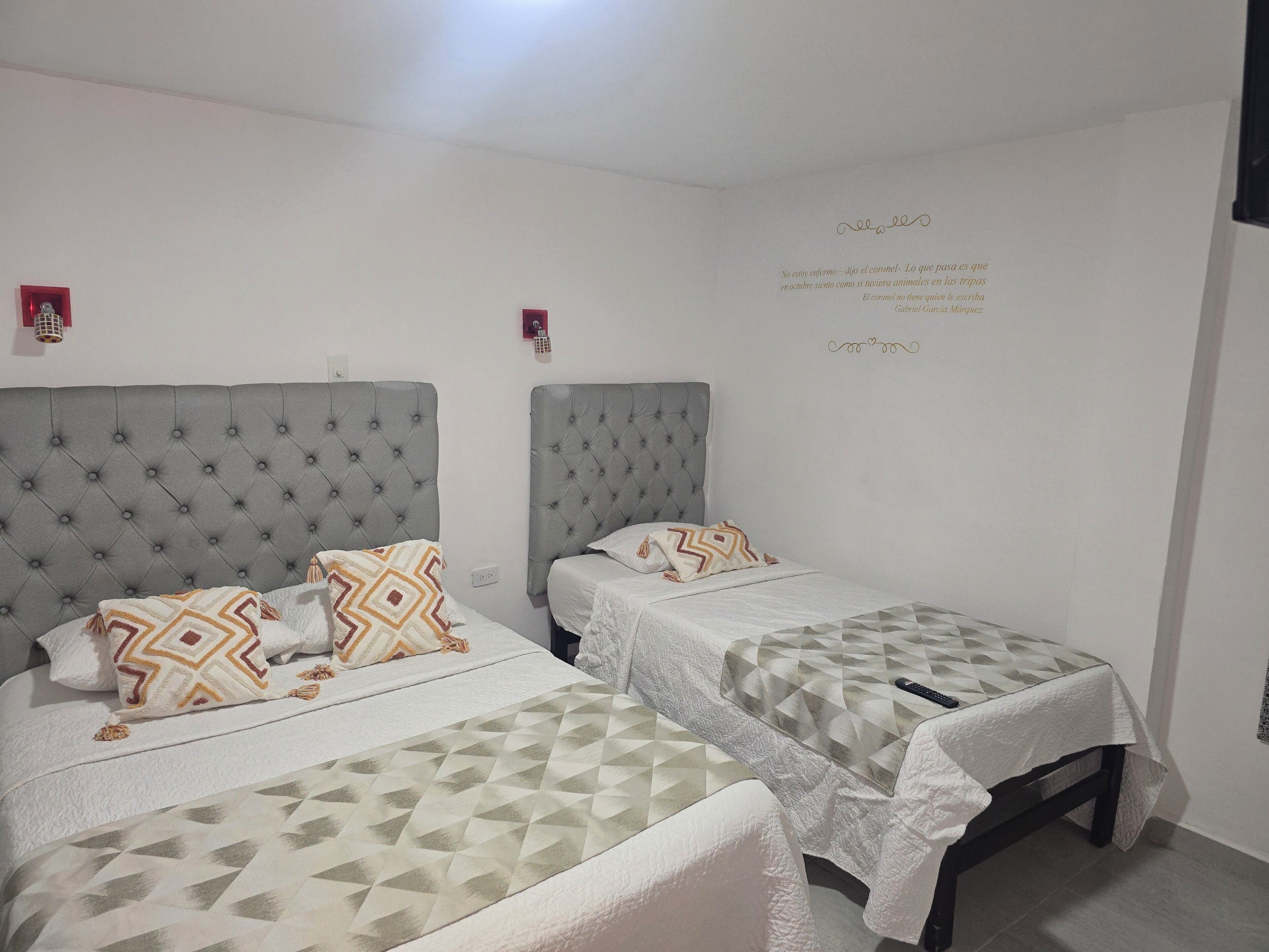 A hotel room with two beds, each with a grey headboard and white bedding. There are decorative pillows and a wall quote in Spanish.