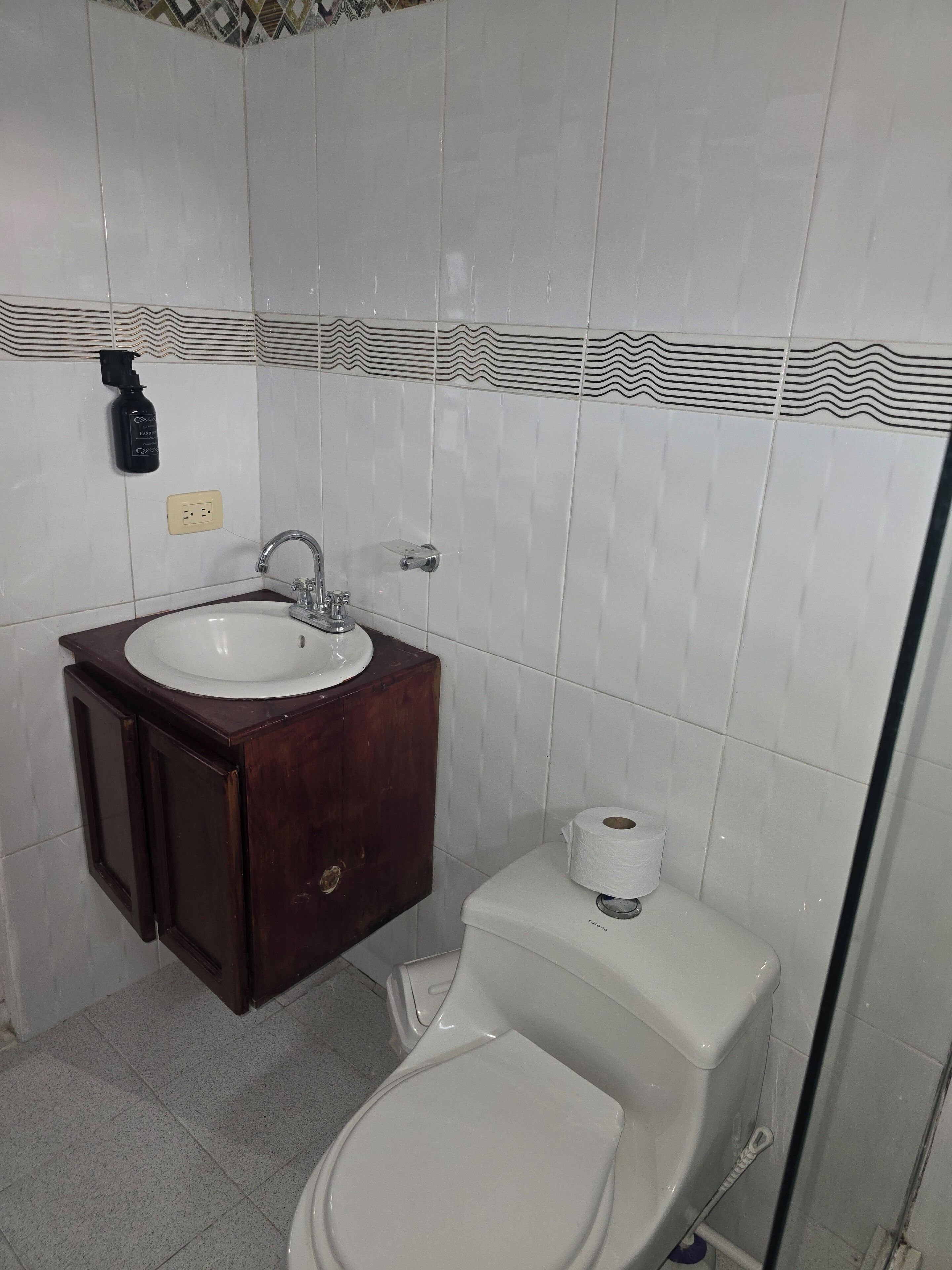 Un baño con lavamanos blanco, tocador de madera oscura, inodoro blanco y baldosas estampadas.