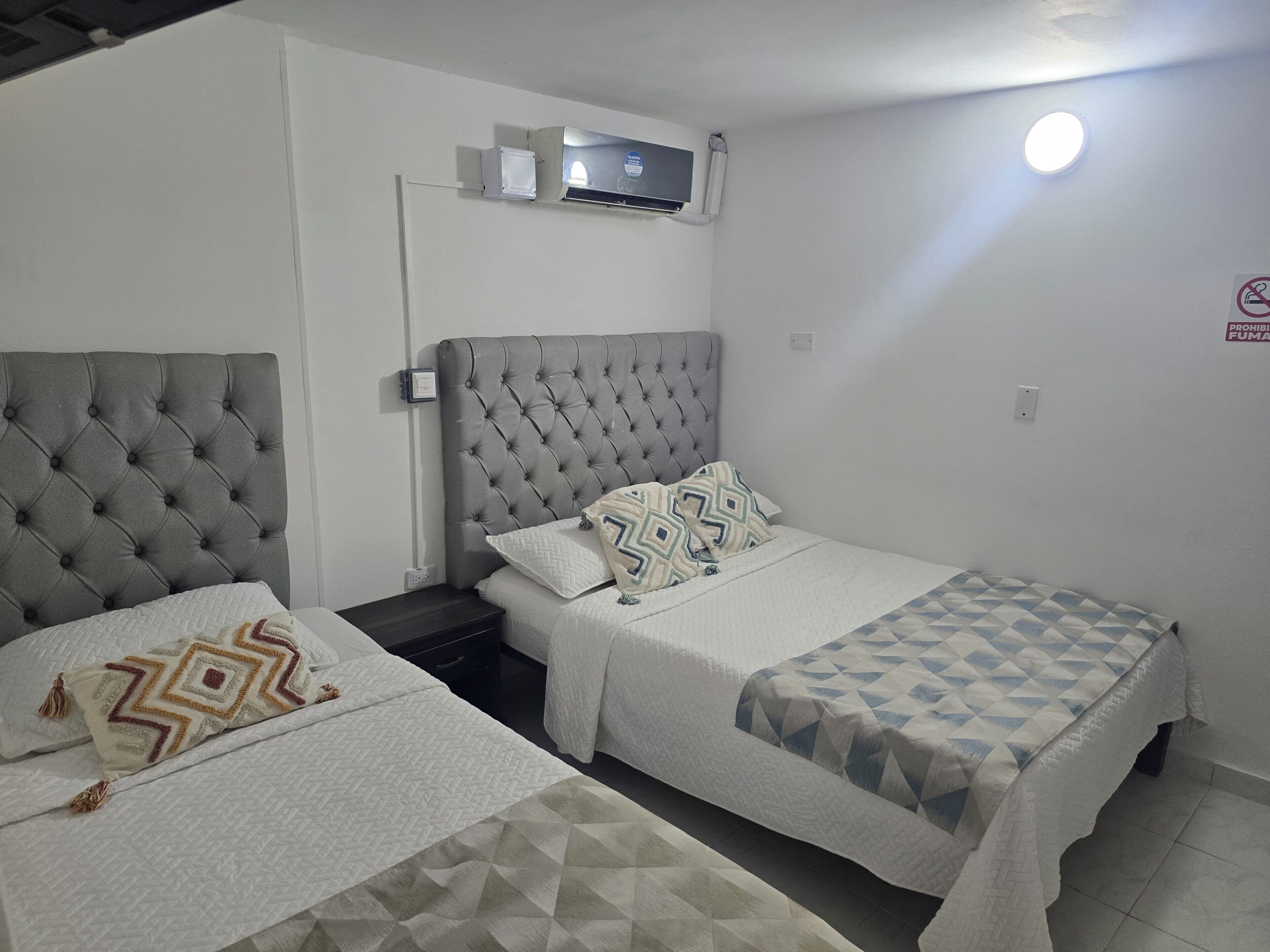 A hotel room with two beds, each with a grey headboard, white bedding, and patterned throw pillows. An air conditioning unit is mounted on the wall above the beds.