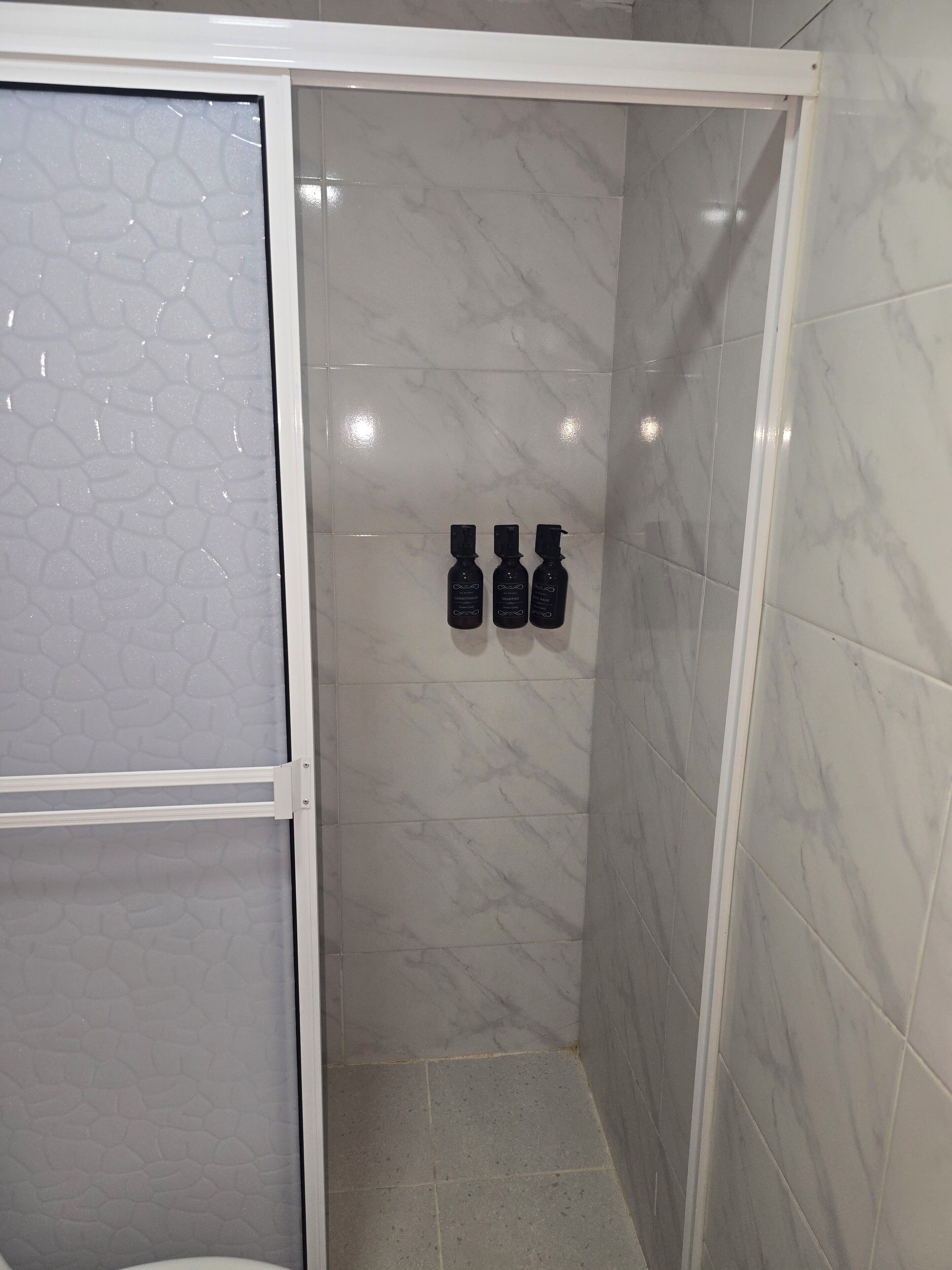 A shower area with white marble tiles, a frosted glass door, and three black bottles on the wall.