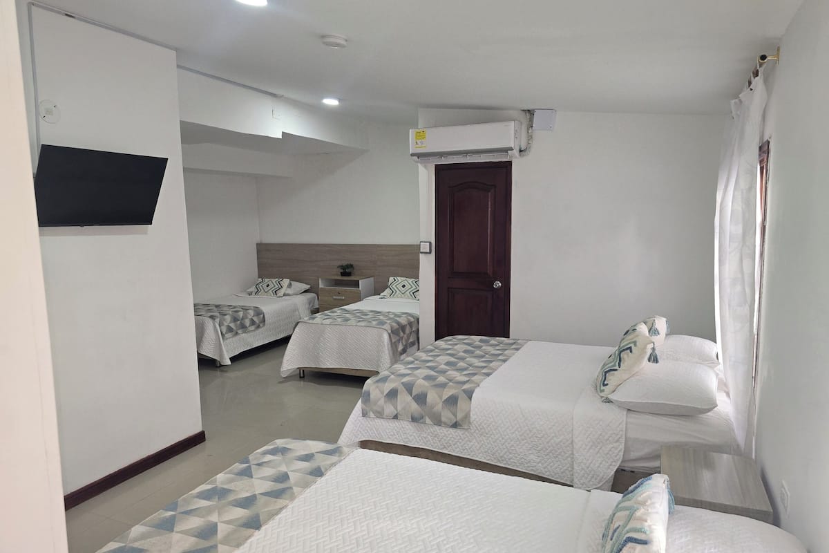 A hotel room with three beds, a wooden headboard, a small bedside table, a TV mounted on the wall, and an air conditioning unit.