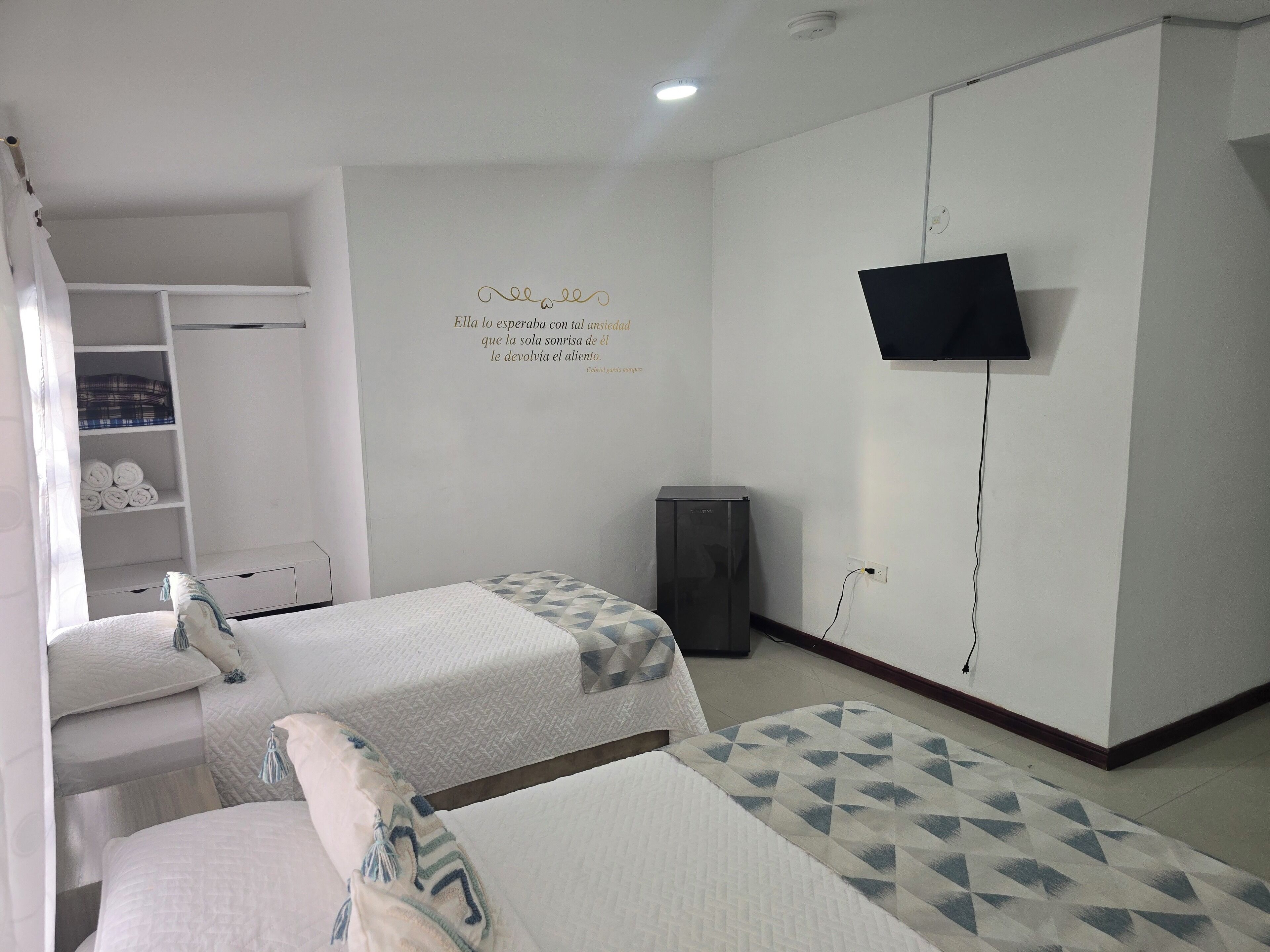 A hotel room with two beds, a wardrobe, a television mounted on the wall, and a shelf with folded towels.