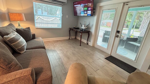 A living room with a grey couch, a TV mounted on the wall, and a window with blinds.