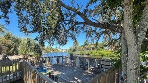 A wooden deck with chairs and tables overlooking a body of water, surrounded by trees and greenery.