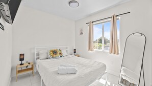 A modern bedroom with a large bed, a bedside table with a lamp, a full-length mirror, and a window with curtains. - Sunflower Miami- A Family Retreat/ Fenced Turf Yard / BBQ-Free Parking (Miami)