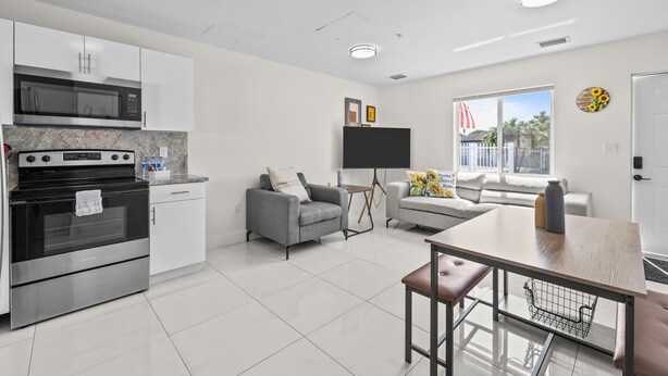 A modern kitchen with stainless steel appliances, a dining area with a wooden table, and a living room with a sofa and a TV. - Sunflower Miami- A Family Retreat/ Fenced Turf Yard / BBQ-Free Parking (Miami)