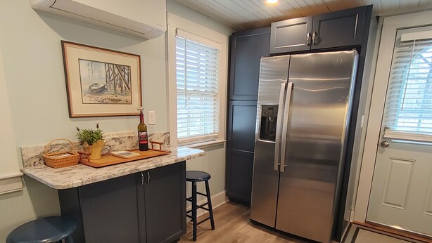 A kitchen with dark cabinets, a stainless steel refrigerator, a marble countertop, a wicker basket, a potted plant, and a framed picture on the wall. - A Dennis Port gem-just steps to the beach! Totally remodeled top to bottom!    (Dennis)