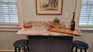 A kitchen with a marble countertop, two bar stools, a wicker basket, a potted plant, a wine bottle, and a framed picture on the wall. - A Dennis Port gem-just steps to the beach! Totally remodeled top to bottom!    (Dennis)