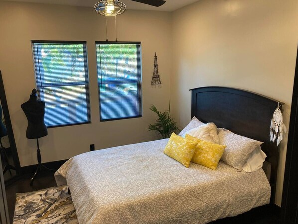 A bedroom with a bed, two windows, a mannequin, a ceiling fan, and a plant.