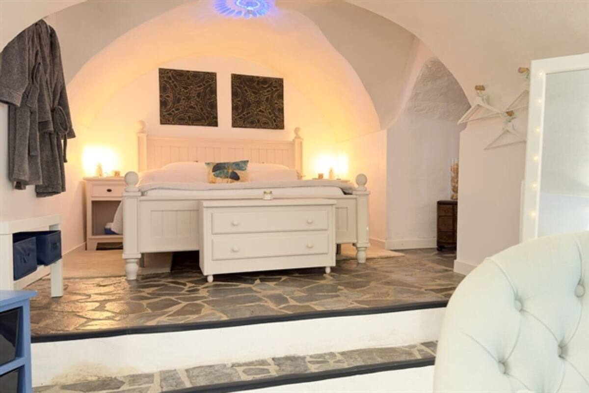 A bedroom with a stone floor, a white bed with two pillows, a white dresser, a robe hanging on the wall, and a white chair.