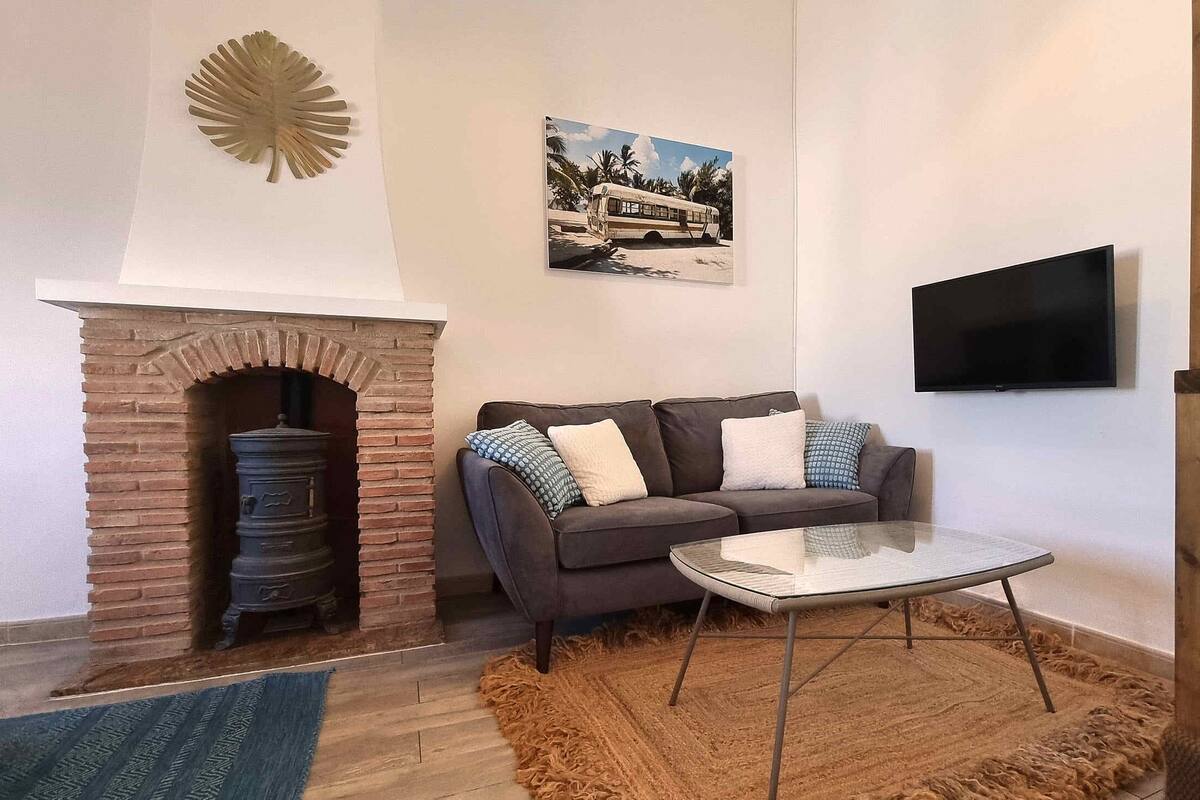 A living room with a brick fireplace, a sofa with patterned cushions, a glass coffee table, a mounted TV, and a wooden staircase.