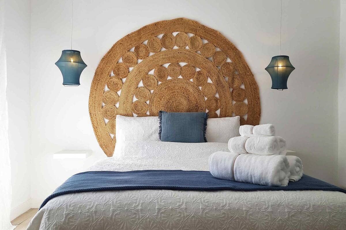 A bed with a woven headboard, two wall-mounted lamps, and a stack of towels.