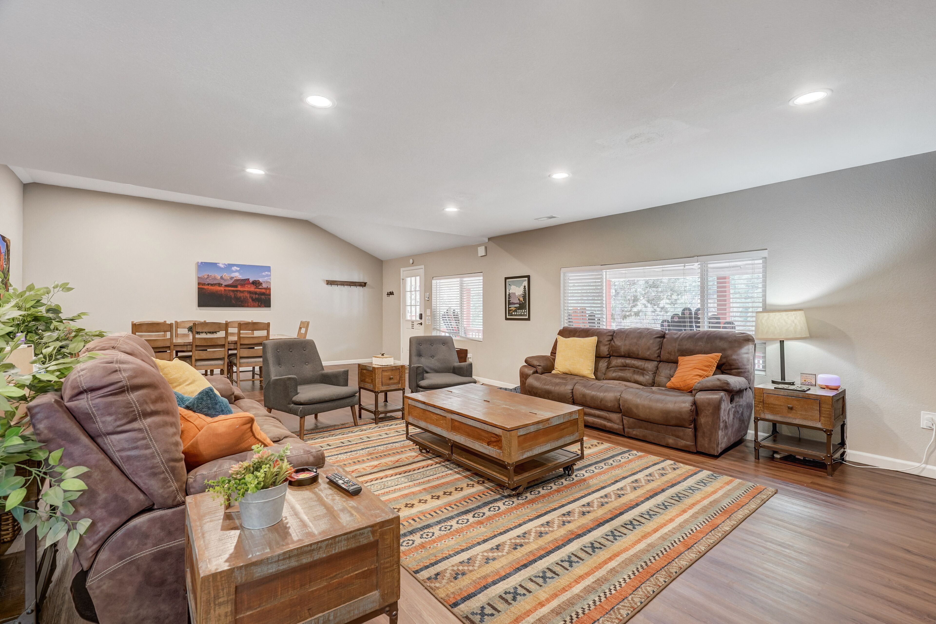 A spacious living room with a brown leather sofa, a wooden coffee table, and a patterned rug. There are two armchairs, a dining table, and a lamp.